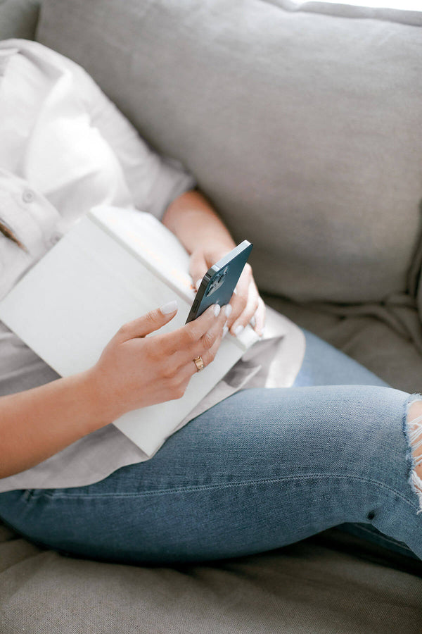 Woman sitting comfortably on a couch with a book and smartphone in hand, browsing resources—symbolizing easy access to trusted pelvic health professionals through the Hem Support Wear directory.